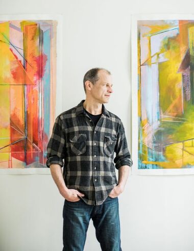 Portrait of Sean Hasey standing with hands in pockets looking to his left. He's between two of his brightly colored large-scale paintings 